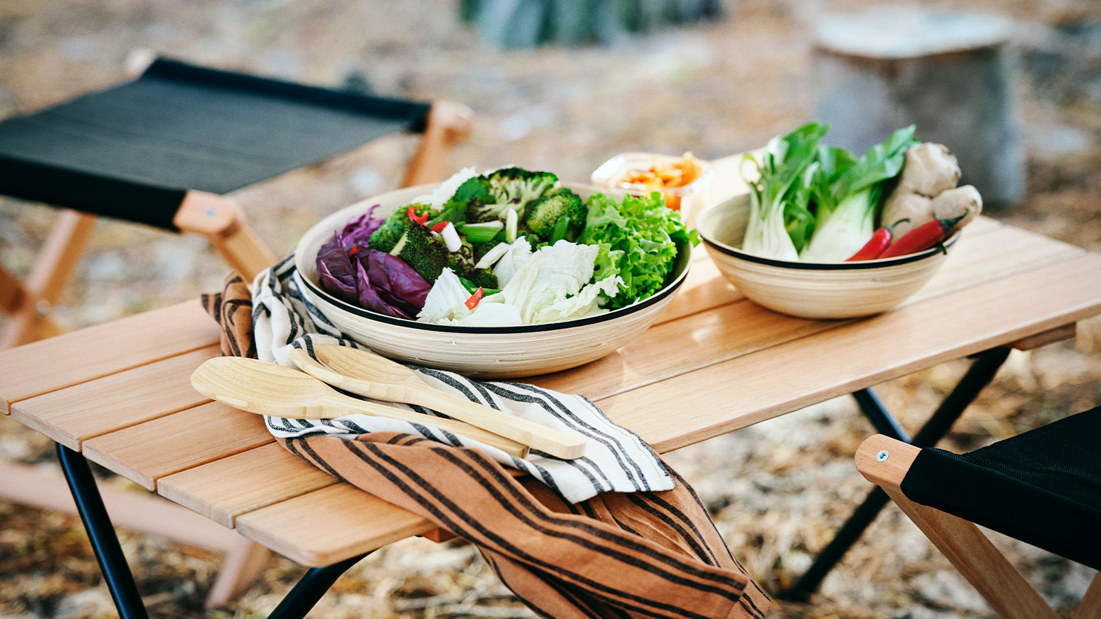 Two large ceramic bowls filled with fresh green salad, broccoli, cabbage, and ginger on a light-colored wooden folding camping table with wooden utensils and striped napkins.