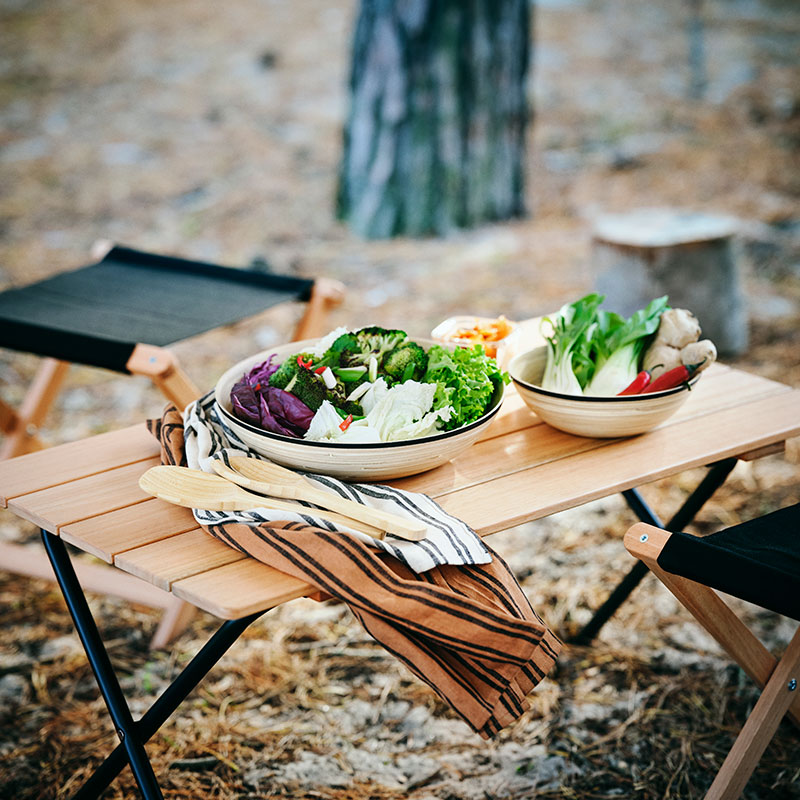 Two large ceramic bowls filled with fresh green salad, broccoli, cabbage, and ginger on a light-colored wooden folding camping table with wooden utensils and striped napkins.