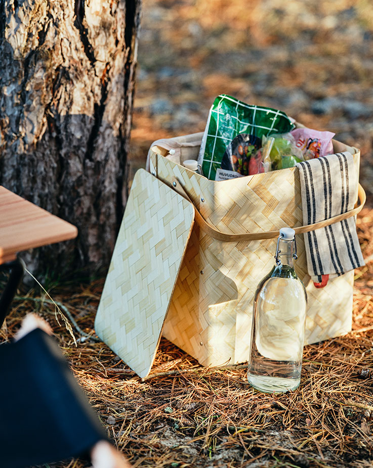 A light-colored woven wooden picnic basket with its lid open, filled with food packages and a striped dish towel, next to a clear glass bottle with a cork on the forest floor.