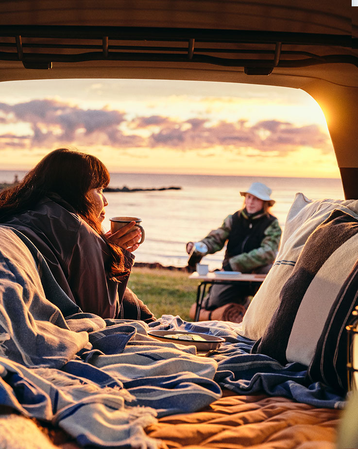 A view from inside the trunk of a car lined with blue blankets and cushions, looking toward a person sitting at a camping table facing the ocean during a warm, golden sunset.
