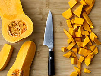 Top-down view of a halved butternut squash, a sleek chef's knife with a black handle, and neatly diced squash pieces arranged on a bamboo cutting board.