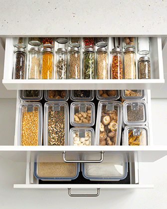 Top-down view of three open white kitchen drawers meticulously organized with glass spice jars and transparent food storage containers filled with dry ingredients.