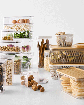 An assortment of clear plastic food storage containers with bamboo lids and mesh baskets stacked on a white surface, holding fresh ingredients and snacks.