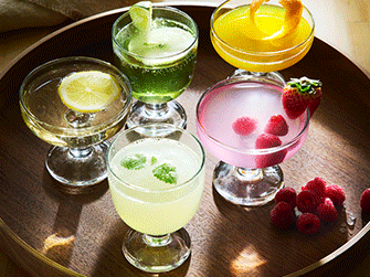 Five elegant glass goblets filled with colorful, garnishes cocktails in yellow, green, and pink, arranged on a dark round wooden tray under warm light.