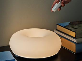 A round donut-shaped table lamp glowing with warm light, next to books, with a hand holding a small remote control above.