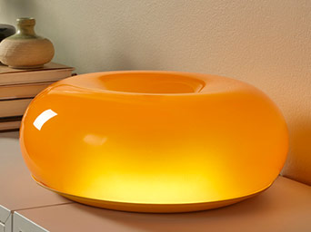 An orange, glossy donut-shaped table lamp glowing softly from within, placed on a light-colored surface.