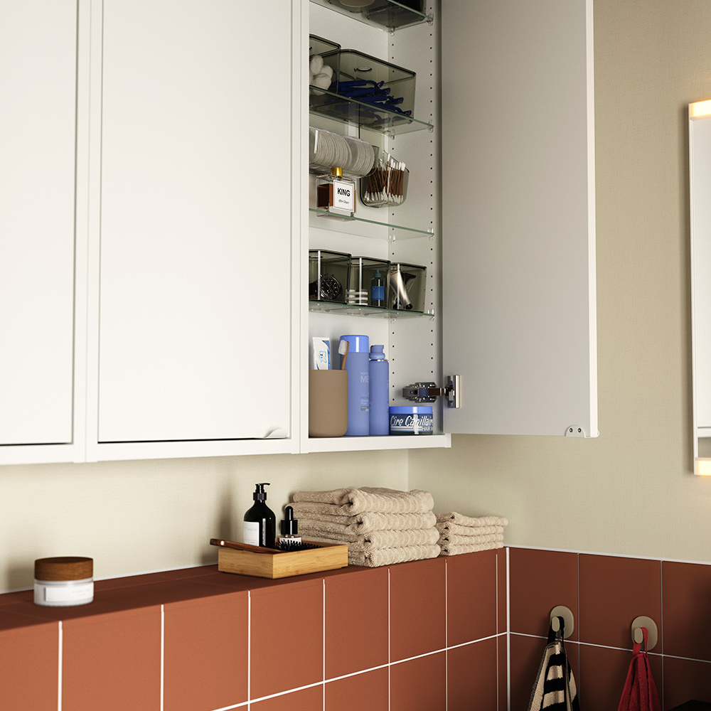 Un baño con azulejos de terracota que cuenta con un armario de pared HAVBÄCK blanco, con una puerta abierta que muestra estantes de vidrio donde se guardan artículos de aseo. 