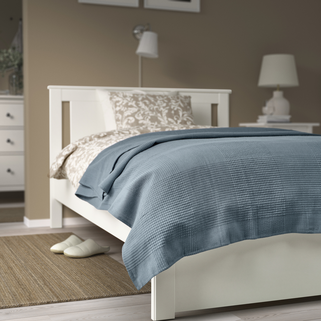 Colcha azul de IKEA sobre cama blanca, almohada beige estampada, lámparas, mesita de noche, zapatillas y alfombra.