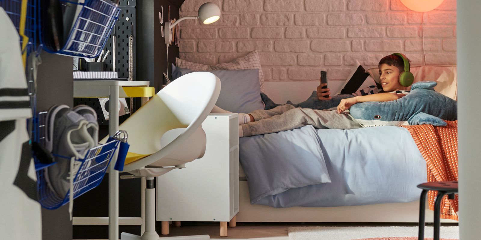 Un adolescente está tumbado en una cama VIHALS escuchando música con los auriculares puestos. En primer plano: un escritorio blanco ENKROKEN y una silla.