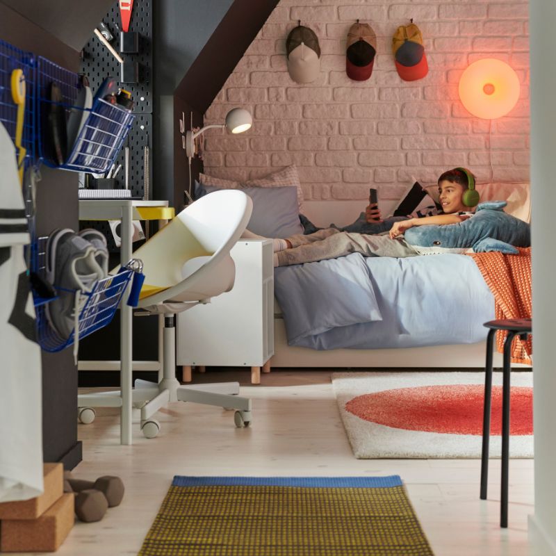 Un adolescente está tumbado en una cama VIHALS escuchando música con los auriculares puestos. En primer plano: un escritorio blanco ENKROKEN y una silla.