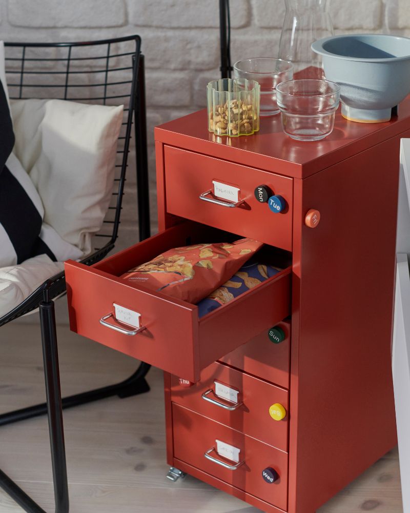 Una habitación con una cómoda roja HELMER con ruedas, con cuatro cajones llenos de aperitivos, junto a una silla moderna.
