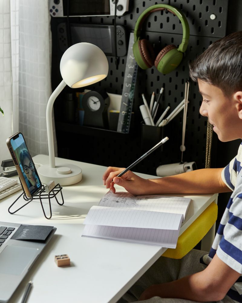 Un niño está sentado ante un escritorio blanco ENKROKEN, con un ordenador portátil, un cuaderno y una lámpara LED blanca KORVSNÖRE encima. 