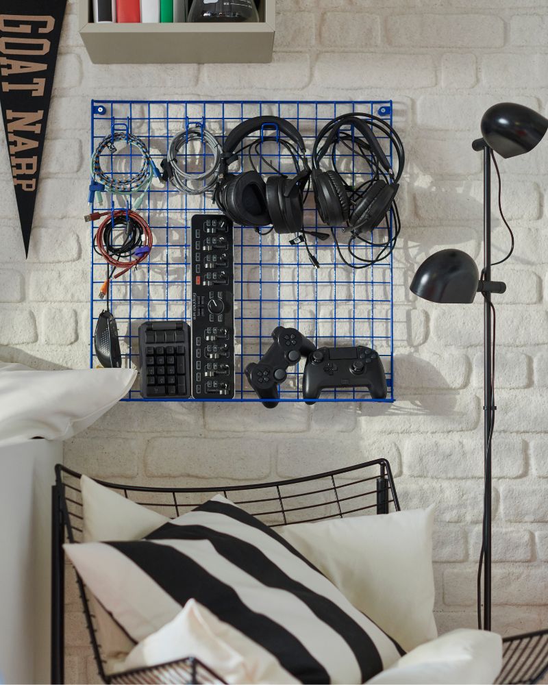 Una rejilla de pared GREJIG azul está colocada en una pared de ladrillo blanco, en la que se guardan auriculares, teclados y mandos de videojuegos.