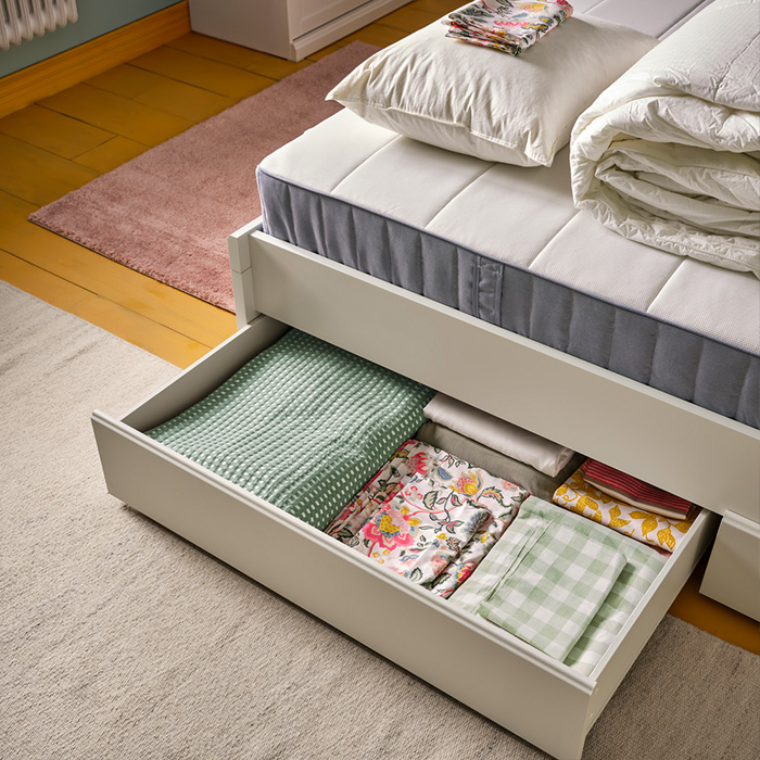 Cama blanca con cajón abierto que guarda ropa de cama colorida, colchón, edredón, almohada y alfombras en el suelo.