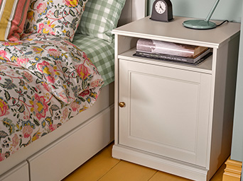 Mesita de noche blanca con libros, lámpara y reloj, junto a una cama con ropa de cama floral y de cuadros.