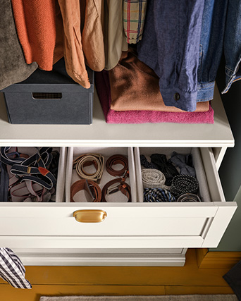 Cómoda blanca con un cajón abierto que organiza cinturones, debajo de ropa colgada, cajas y prendas dobladas.