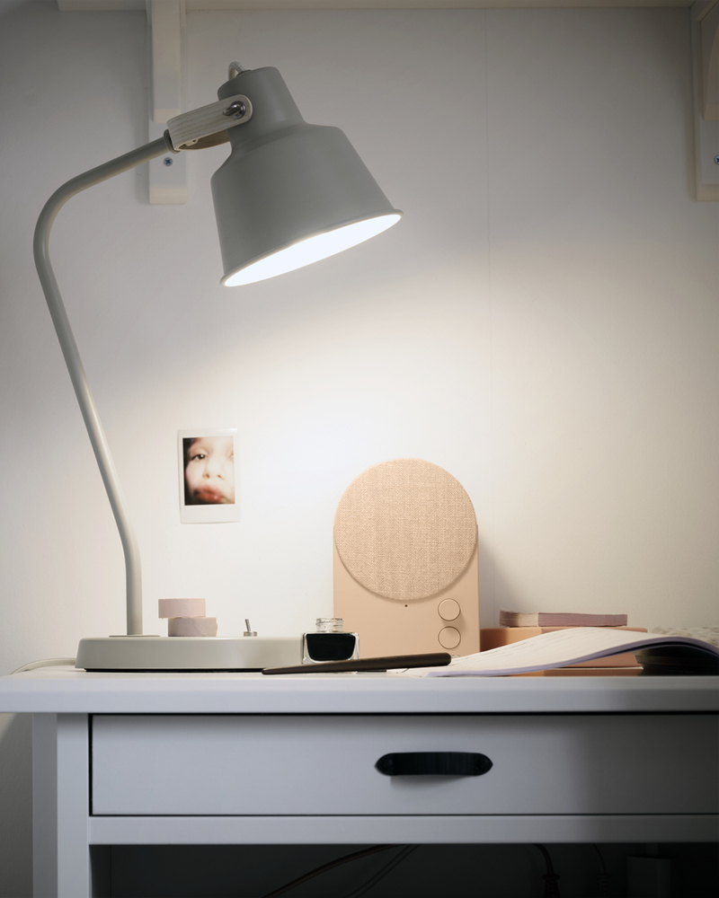 A study area features a beige SJÖMAKT desk lamp made from ash, illuminating a small table beside a photo and stationery.