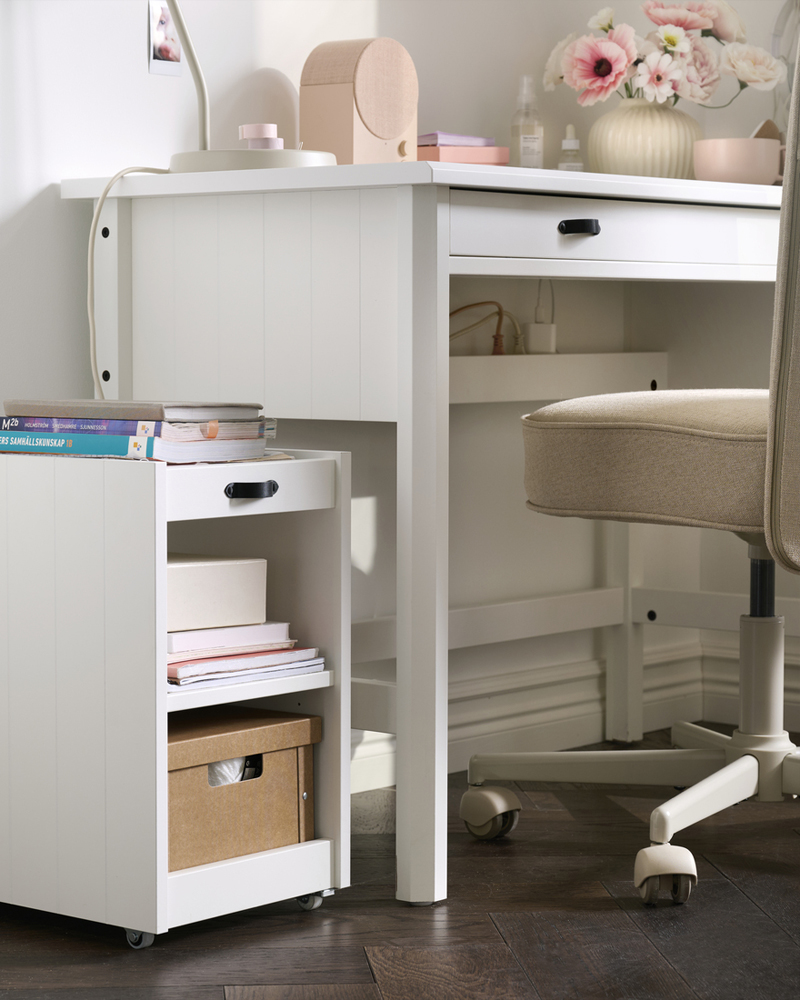 A home office features a white BRYTNING storage unit next to a desk, holding books and boxes for organisation.