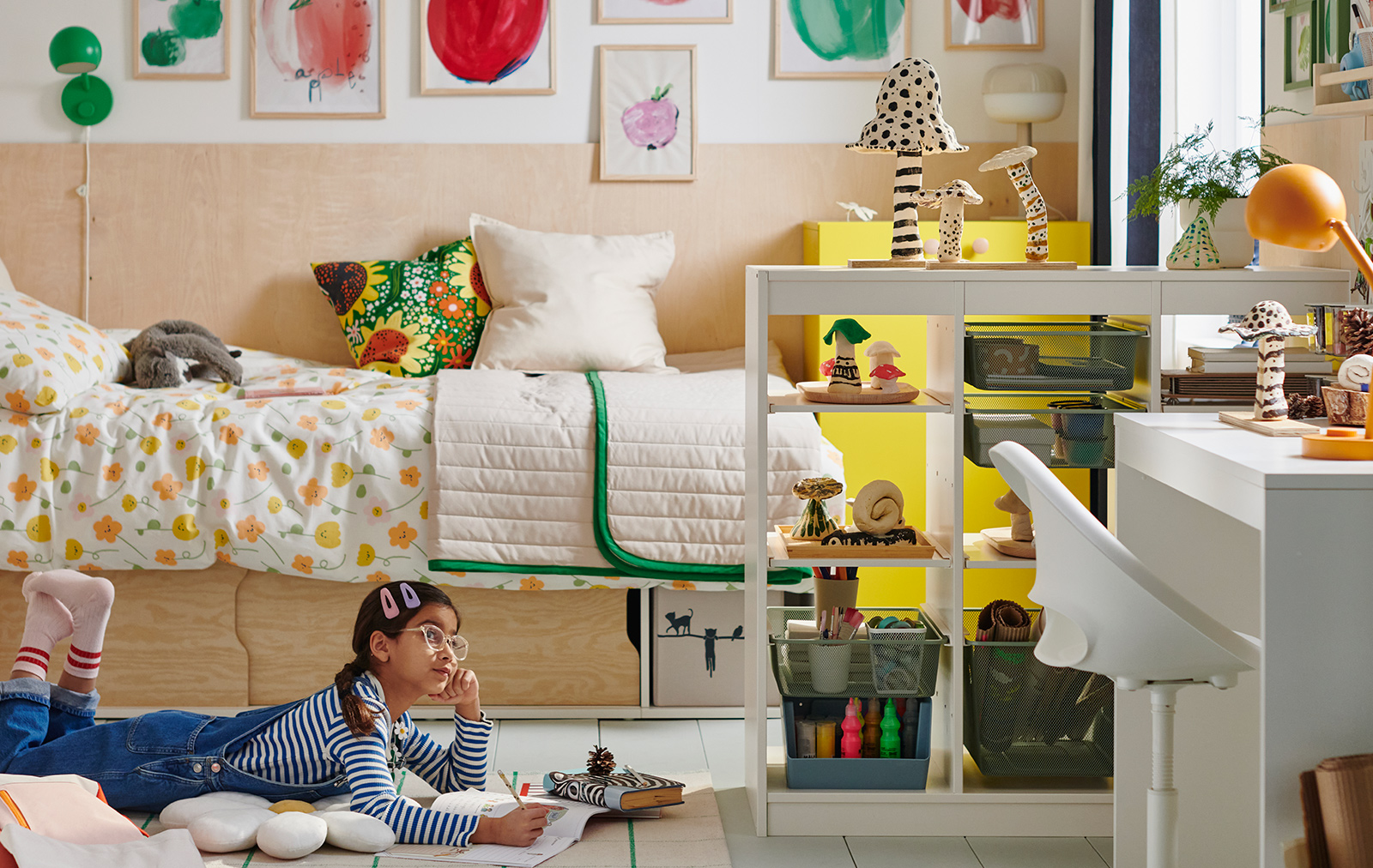 A young girl lies reading on a rug in front of a KLIPPUGGLA bed frame with storage, dressed in floral bedding and pillows.