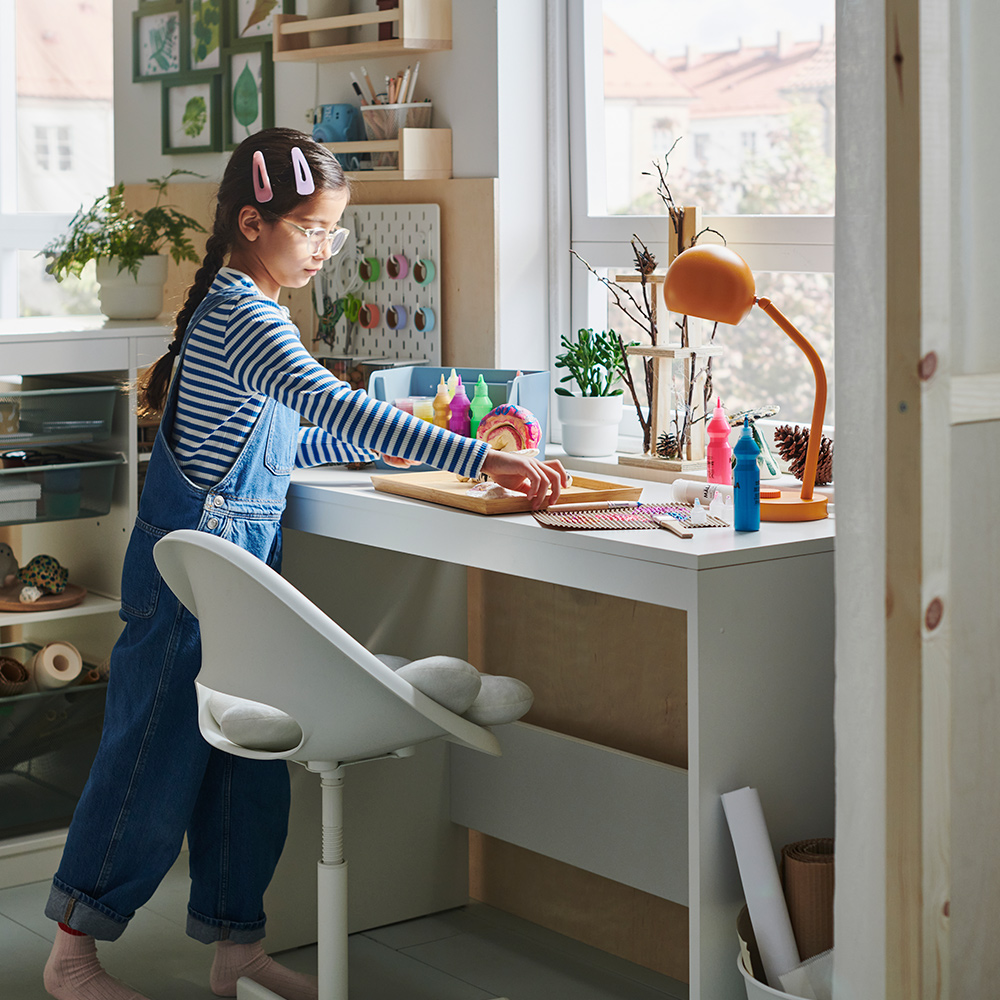 A young girl stands at a white LASTARE desk while lifting an OSTBIT bamboo tray featuring her latest hobby project: a snail.