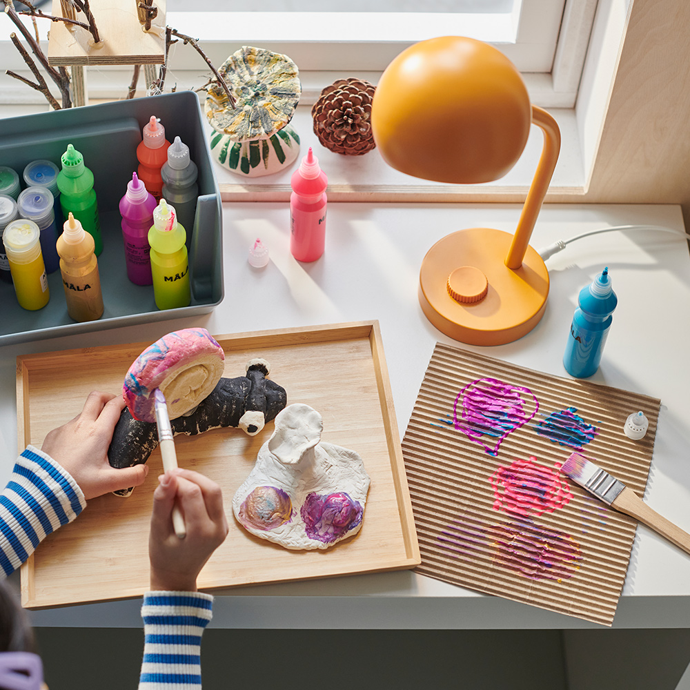 A bright room with a small desk with a yellow KORVSNÖRE lamp and paint bottles. A child paints a snail on an OSTBIT tray.