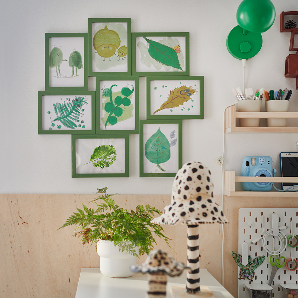 A room with a green VÄXBO collage frame for eight drawings arranged on a white wall above plant-filled shelves.