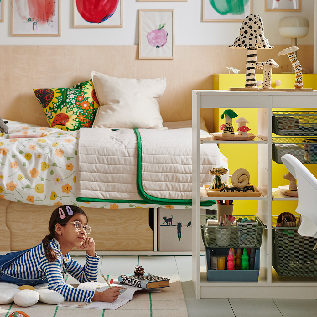 A young girl lies reading on a rug in front of a KLIPPUGGLA bed frame with storage, dressed in floral bedding and pillows.