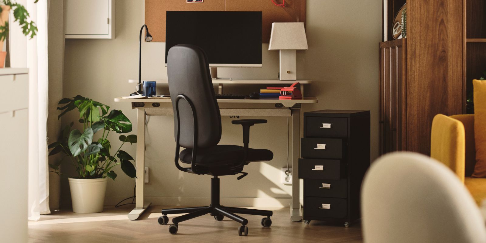An office with a dark grey SMÖRKULL office chair with armrests beside a desk and plants near a wall.