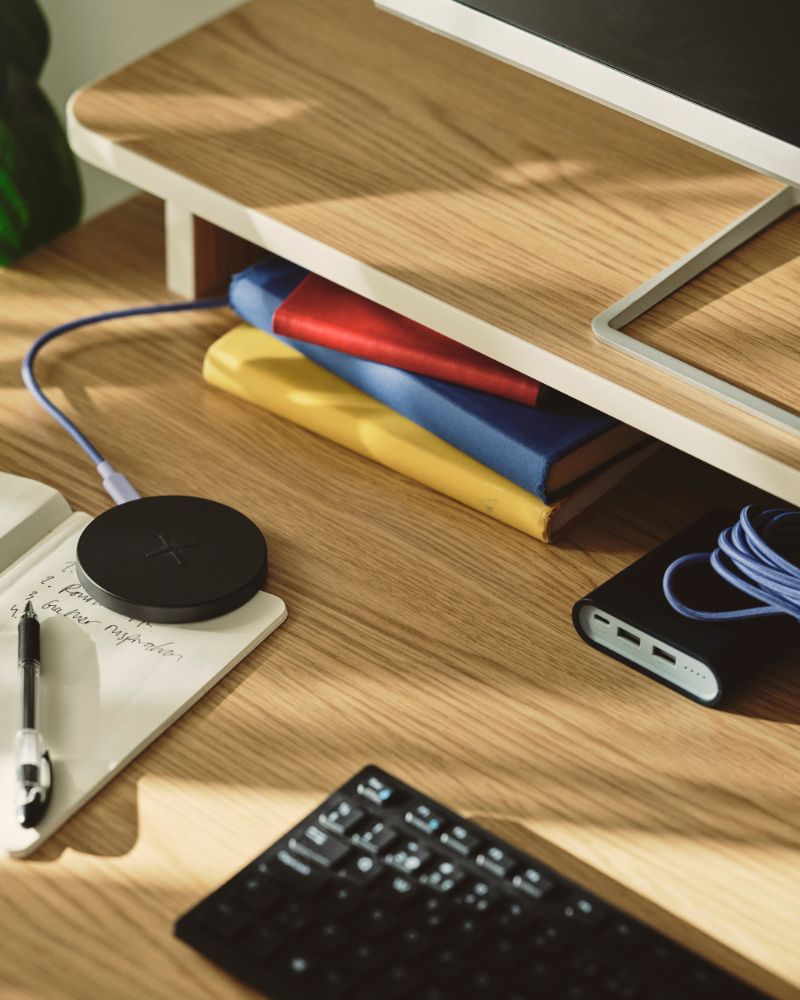 A close-up of a black LIVBOJ wireless charger on a desk, with a notebook, pen, and colourful books nearby.