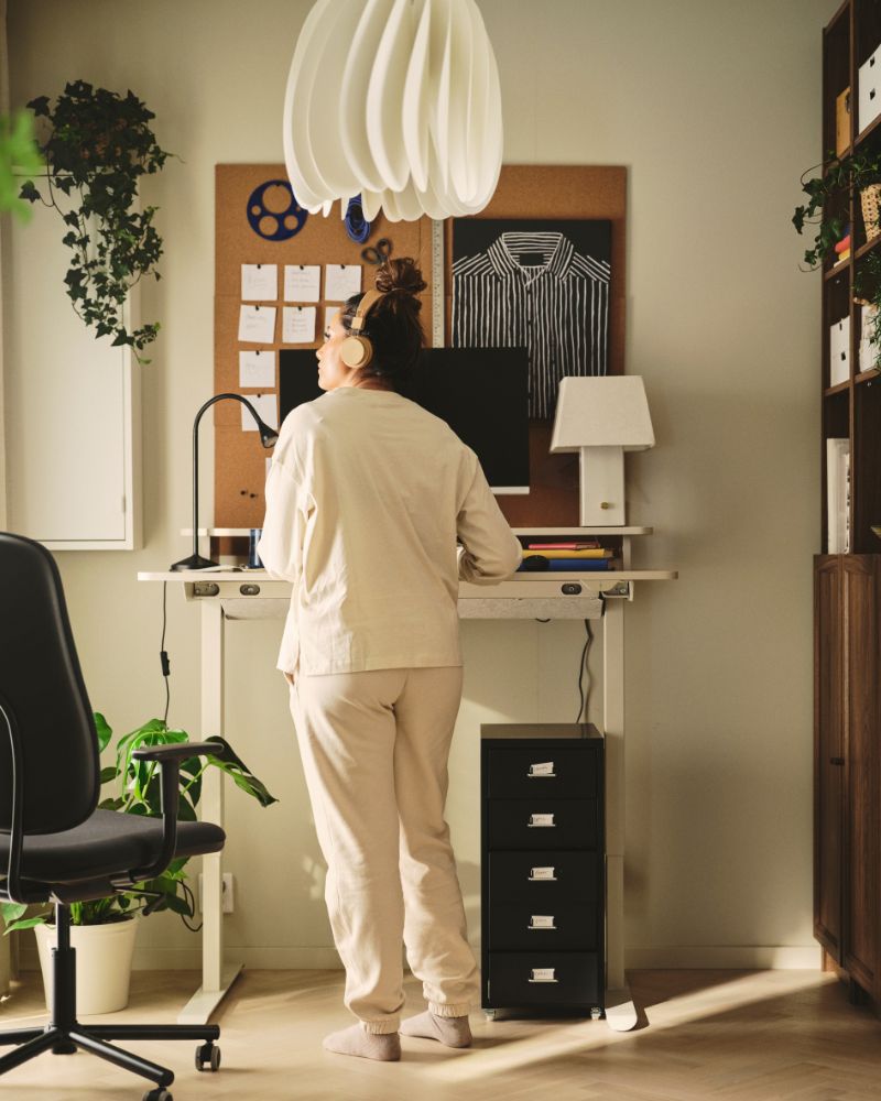 A home office with a beige TRAVSPORT desk sit/stand in oak effect, next to a black rolling chair and potted plants.