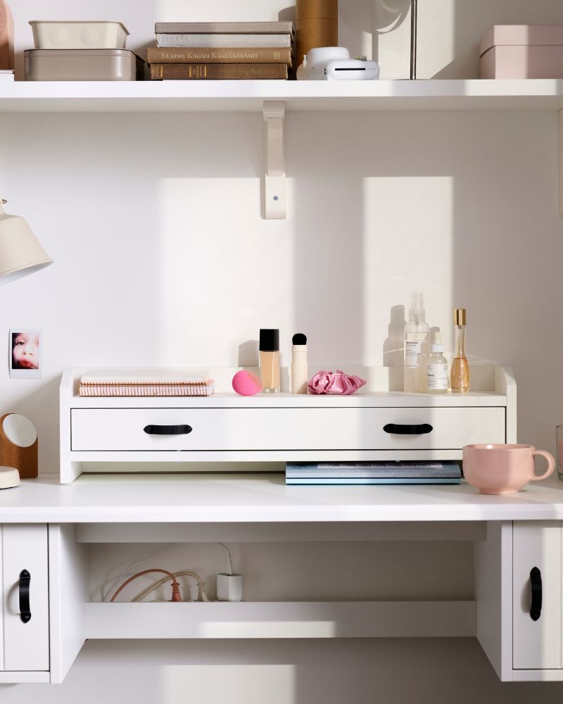 A workspace with a white BRYTNING add-on unit holds stationery and décor, alongside a white lamp and a pink tray.