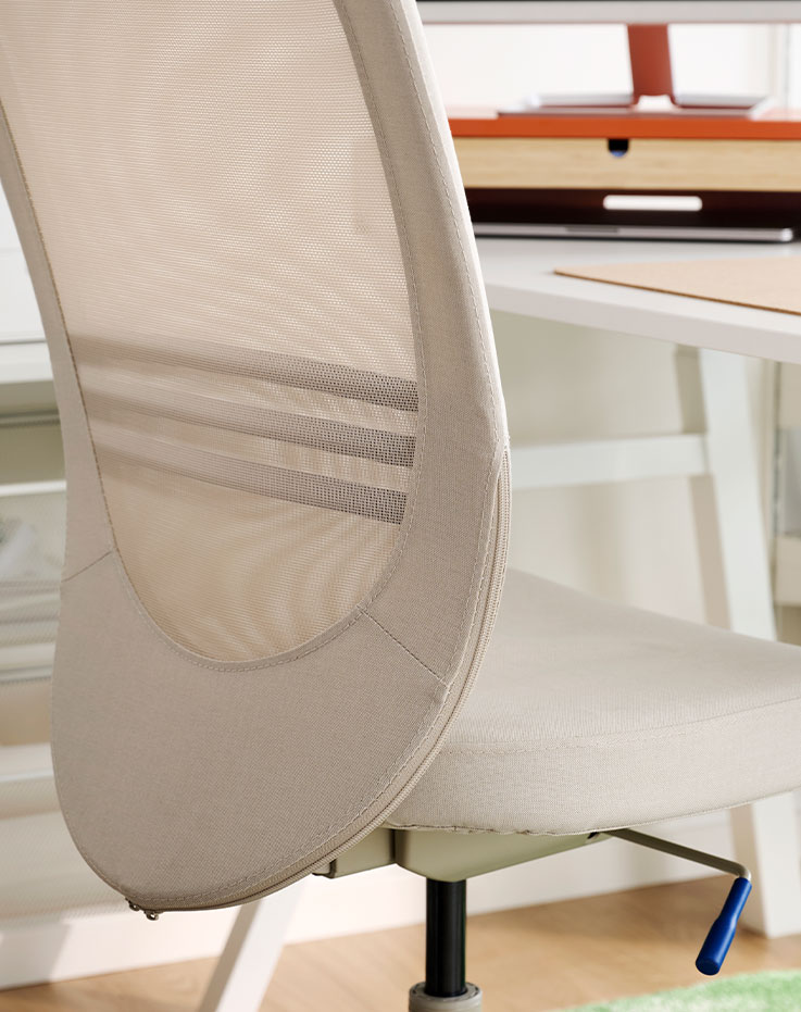 Se ve un primer plano de una silla de oficina FLINTAN de color beige con respaldo de malla, situada en un luminoso espacio de oficinas junto a un escritorio.