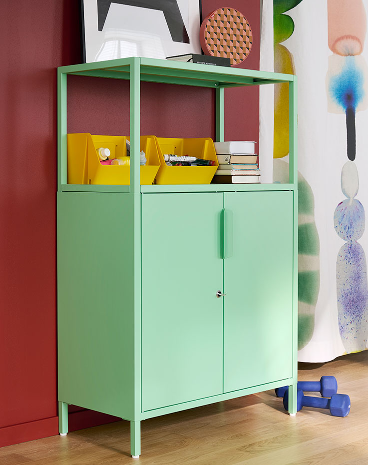 En una habitación luminosa destaca un armario TROTTEN de color verde claro con puertas, en el que se guardan materiales y libros junto a unos coloridos cuadros colgados en la pared.