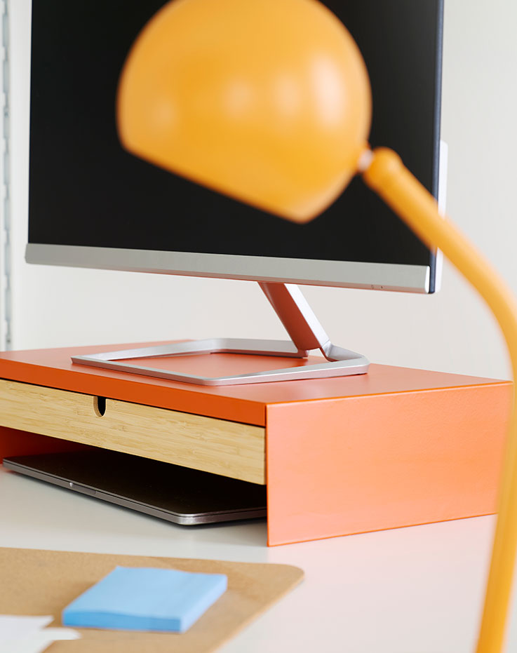 Una vista de primer plano de un soporte para monitor ELLOVEN de color naranja brillante con un cajón, colocado sobre un escritorio junto a un ordenador.