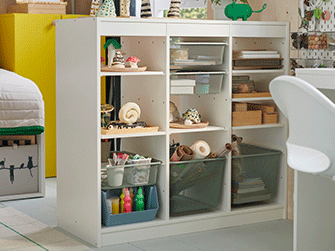 White low shelving unit with three compartments storing craft supplies, paint bottles, and grey mesh storage baskets in a bright room.
