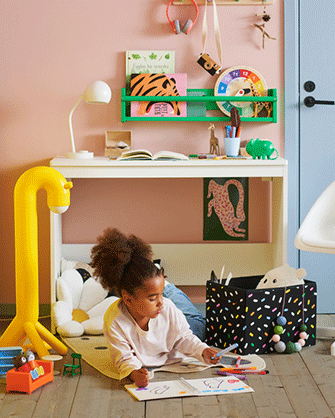 Girl drawing on the floor in front of a white desk with a green bookshelf, yellow floor lamp, and a black patterned storage box.