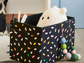 Black fabric storage box with colorful sprinkle pattern filled with a white plush toy, decorated with a string of colorful wooden beads.