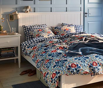 A blue painted bedroom with a floral SÄTERVIDE duvet cover set on a white bed, next to a white bedside table with a lamp.