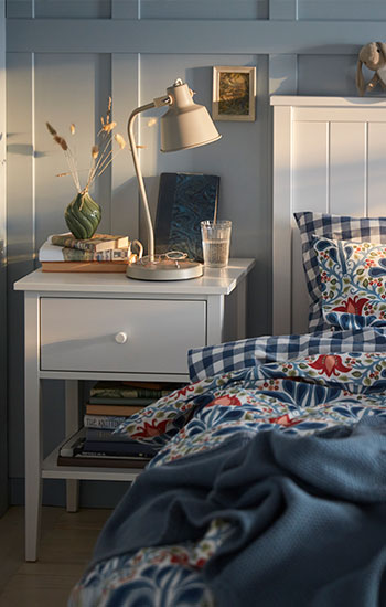 A white bed with a floral multicoloured SÄTERVIDE duvet cover set next to a white bedside table with a lamp.