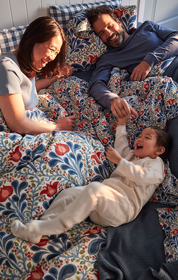 A family laughing in a bed with a floral multicoloured SÄTERVIDE duvet cover set in orange and blue.