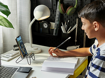 Boy writing in a notebook at a white desk while video calling on a smartphone held by a wire stand.