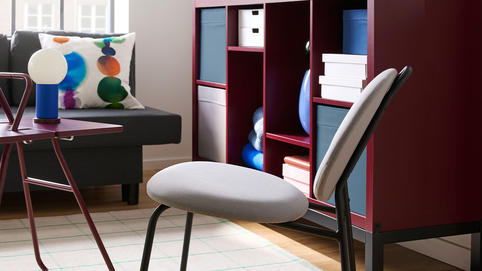 A living room with a brown-red/black KALLAX shelving unit with underframe holds books and decorative items next to a chair.
