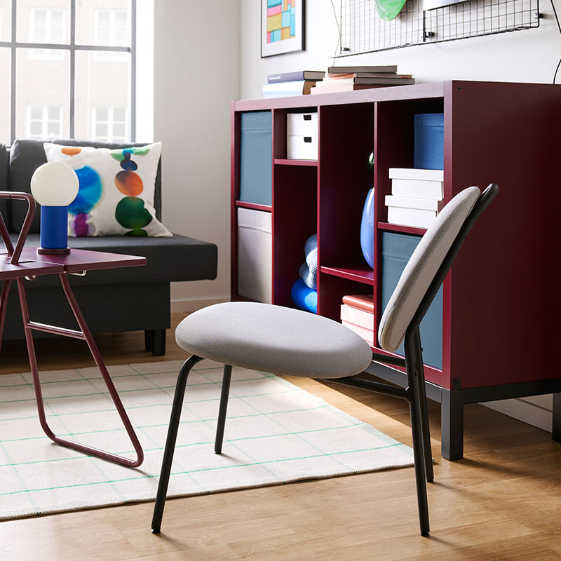 A living room with a brown-red/black KALLAX shelving unit with underframe holds books and decorative items next to a chair.