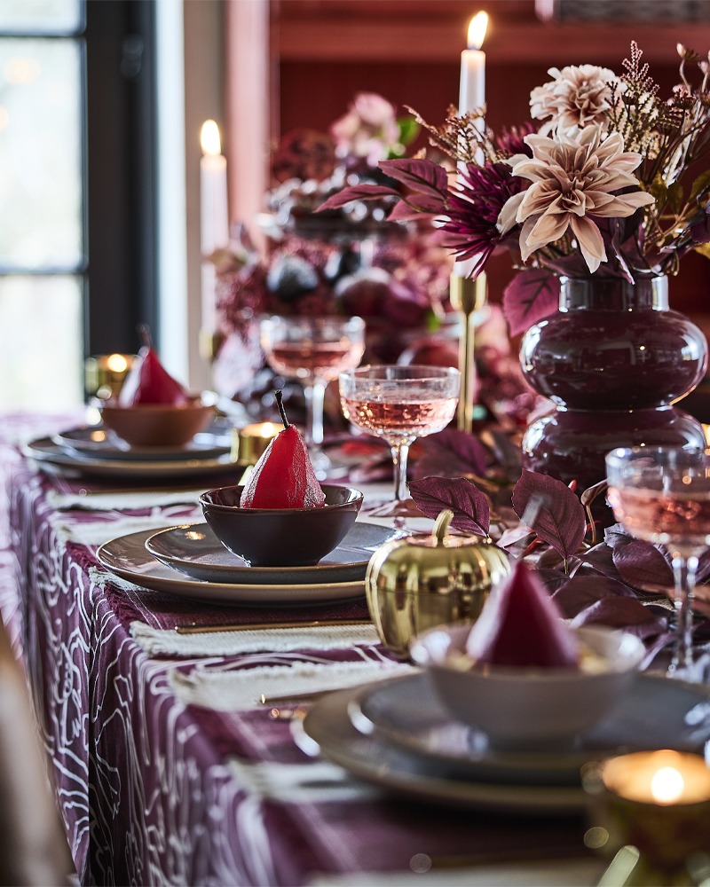 Una mesa decorada en tonos rosa y rojo con velas encendidas, ramos artificiales de HÖSTAGILLE en jarrones y peras escalfadas en cuencos.