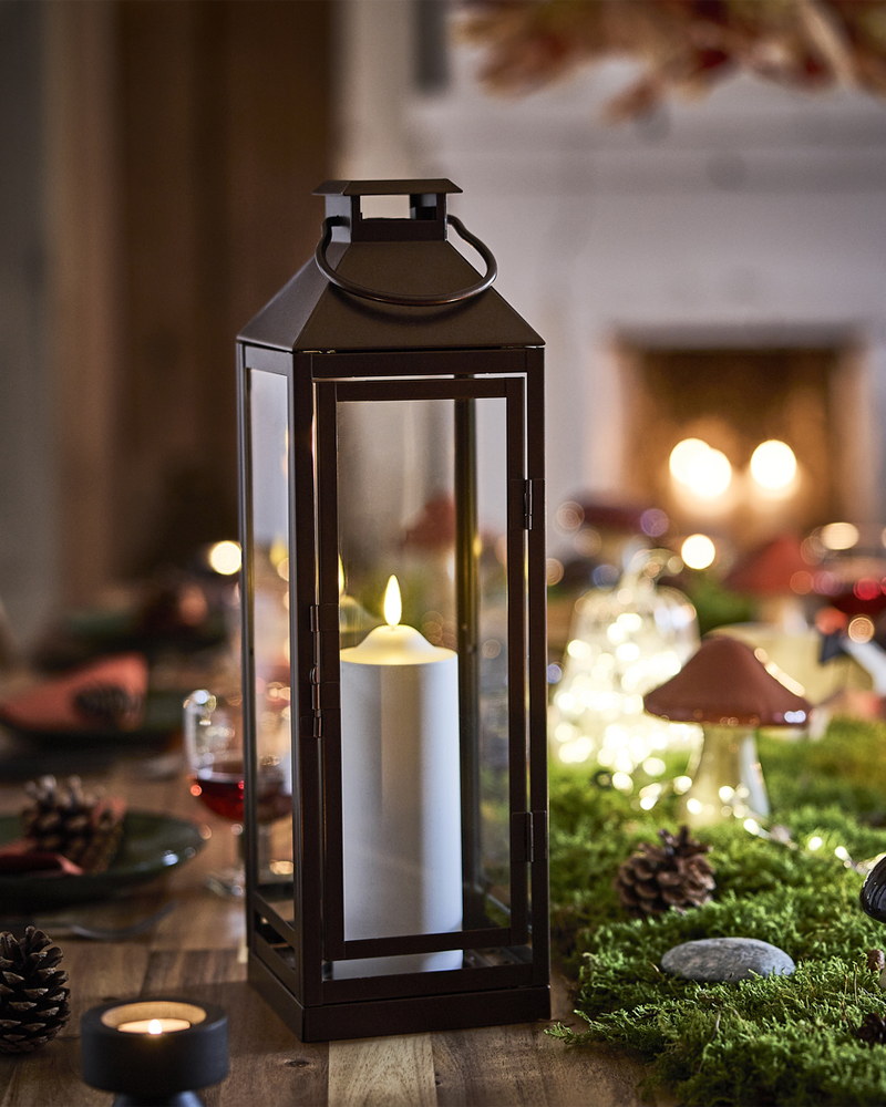 Una mesa decorada con temática forestal, con una vela encendida dentro de un farol junto a setas decorativas sobre musgo artificial.