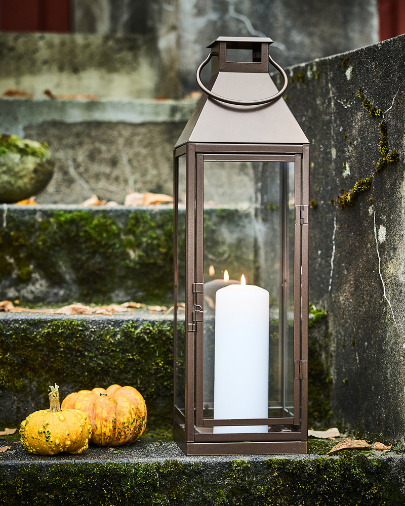 Una vela blanca de columna encendida dentro de un farolillo de HÖSTAGILLE junto a dos mini calabazas en una escalera exterior.