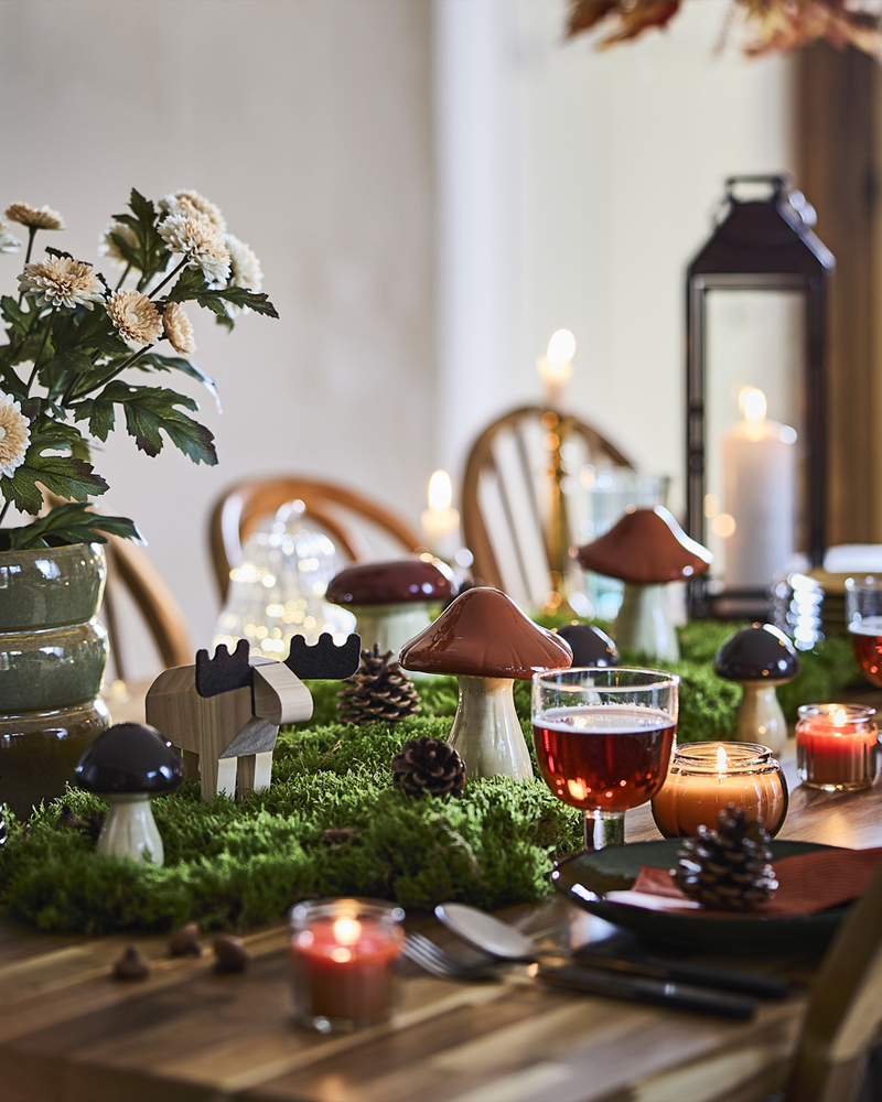 Una mesa de comedor con temática forestal está decorada con setas y alces decorativos de la colección HÖSTAGILLE, velas encendidas, musgo y piñas.
