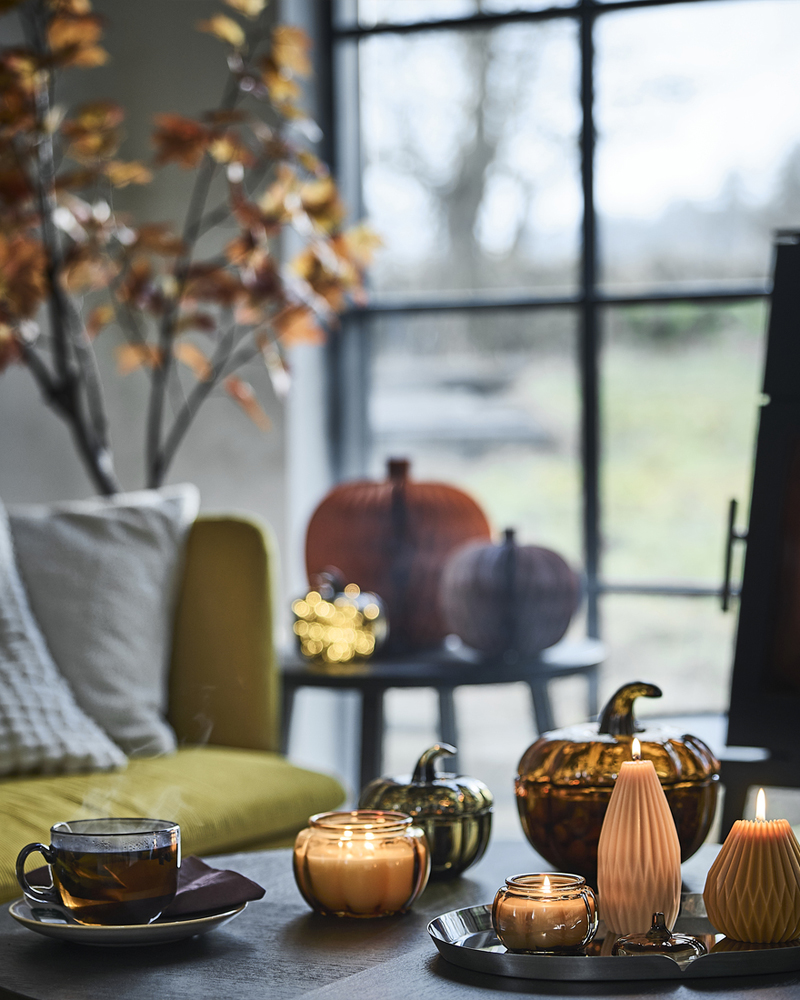 En un rincón acogedor junto a una ventana hay una mesita con velas perfumadas HÖSTAGILLE encendidas, velas cilíndricas con forma de calabaza y una taza.