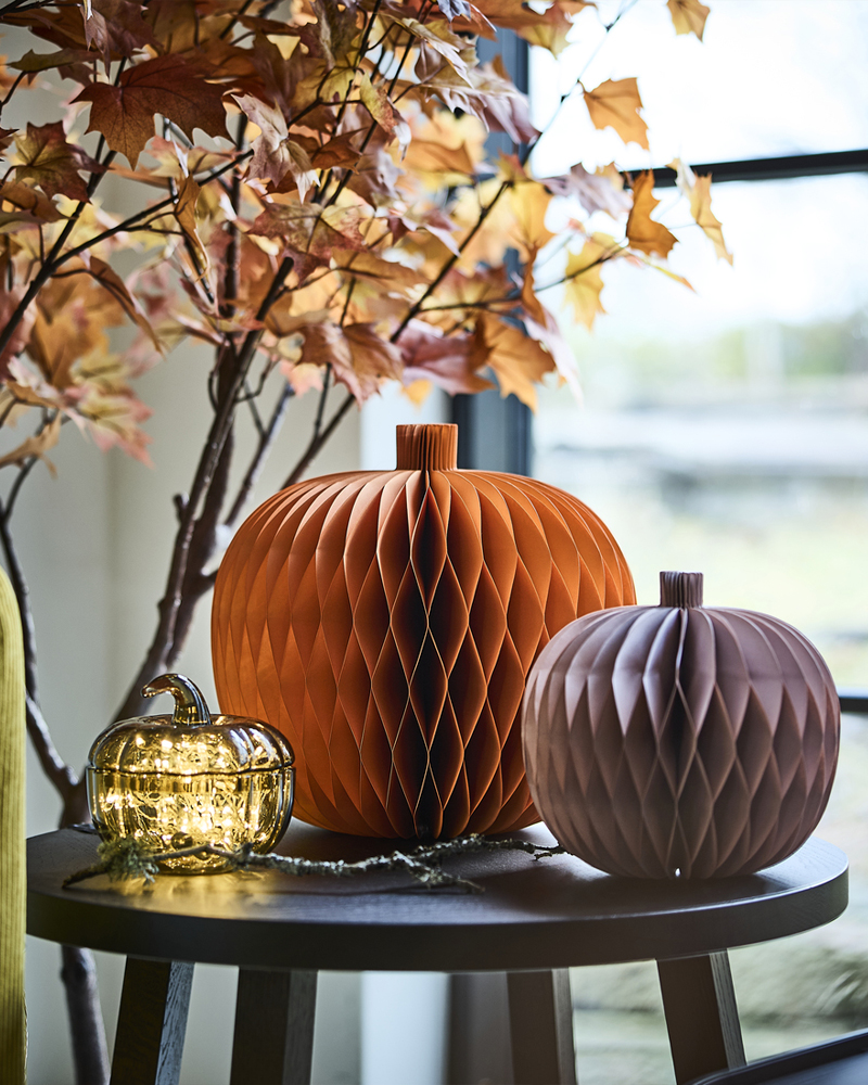 Un juego de dos calabazas de papel decorativas de HÖSTAGILLE y un cuenco con forma de calabaza y tapa se encuentran sobre una mesita auxiliar junto a la ventana.
