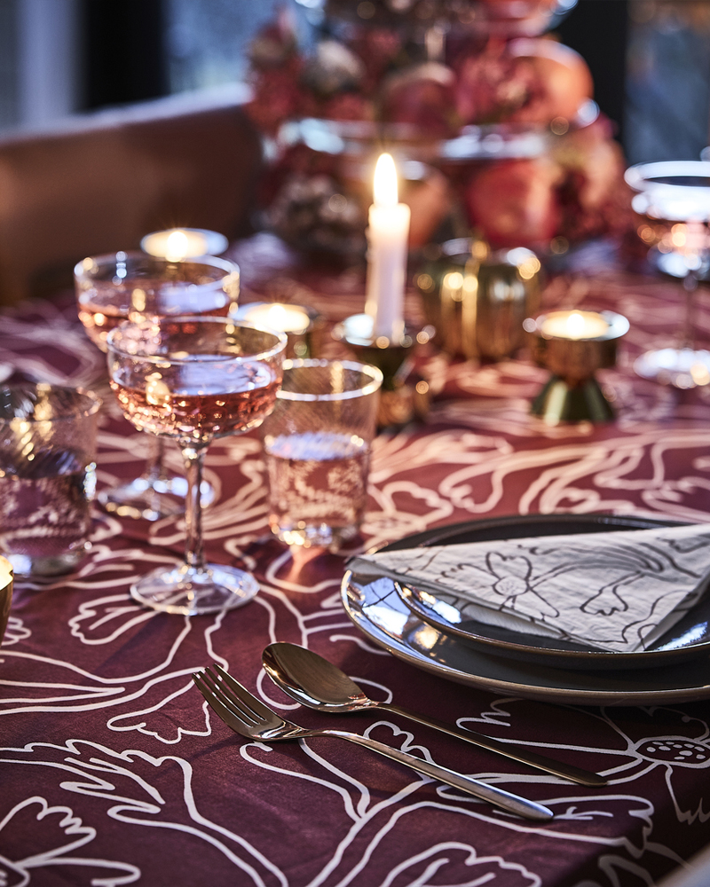 Una mesa de comedor rosa y roja está puesta con velas encendidas, un mantel HÖSTAGILLE con motivos rojos, servilletas de papel y copas.
